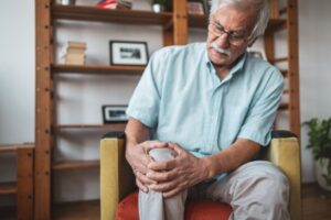 Person seated on a chair indoors, holding one knee with both hands, indicating knee pain