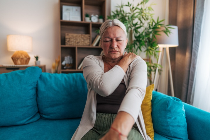 Person seated on a couch holding one shoulder, indicating shoulder or upper‑neck pain